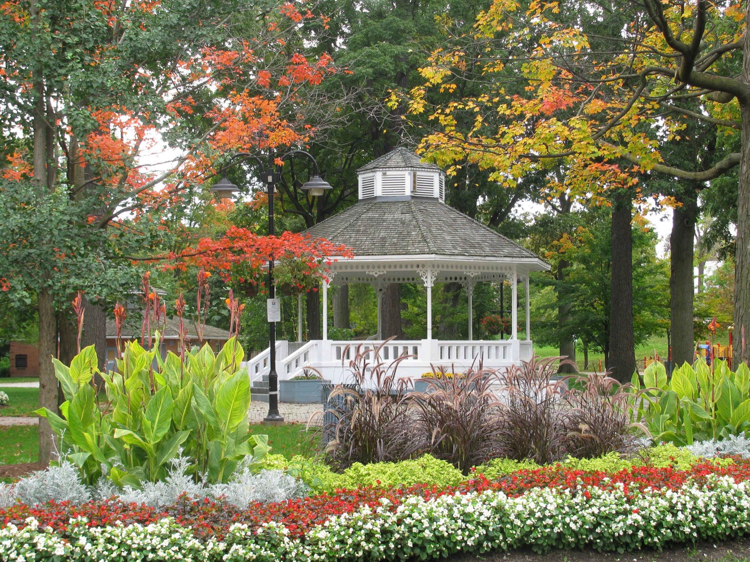 The white gazebo in Gage Park in downtown Brampton, behind beds of red and white flowers and maples turning orange in the autumn