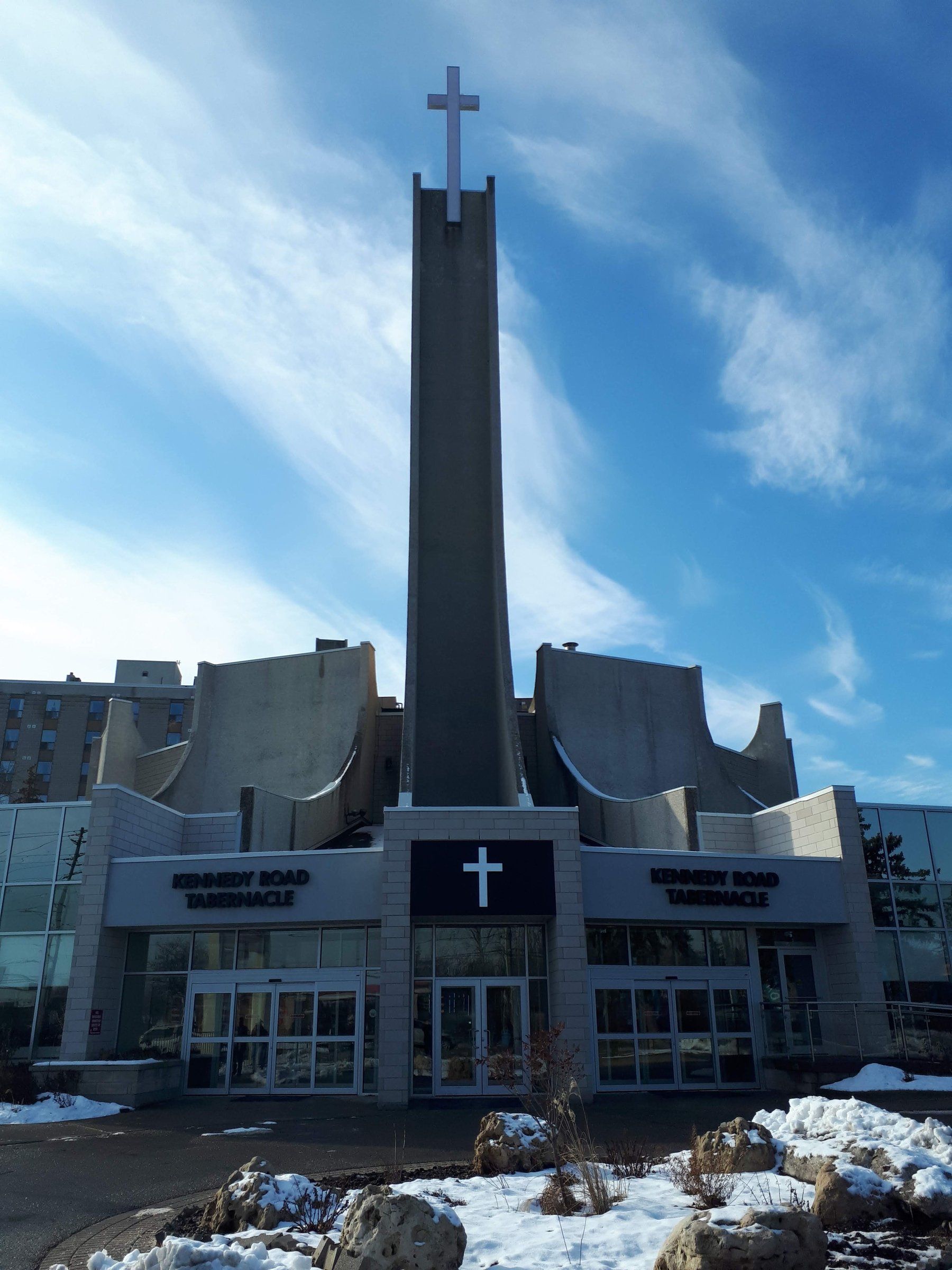 The tall concrete tower and cross above the glass entrance of Kennedy Road Church, with snow on the ground