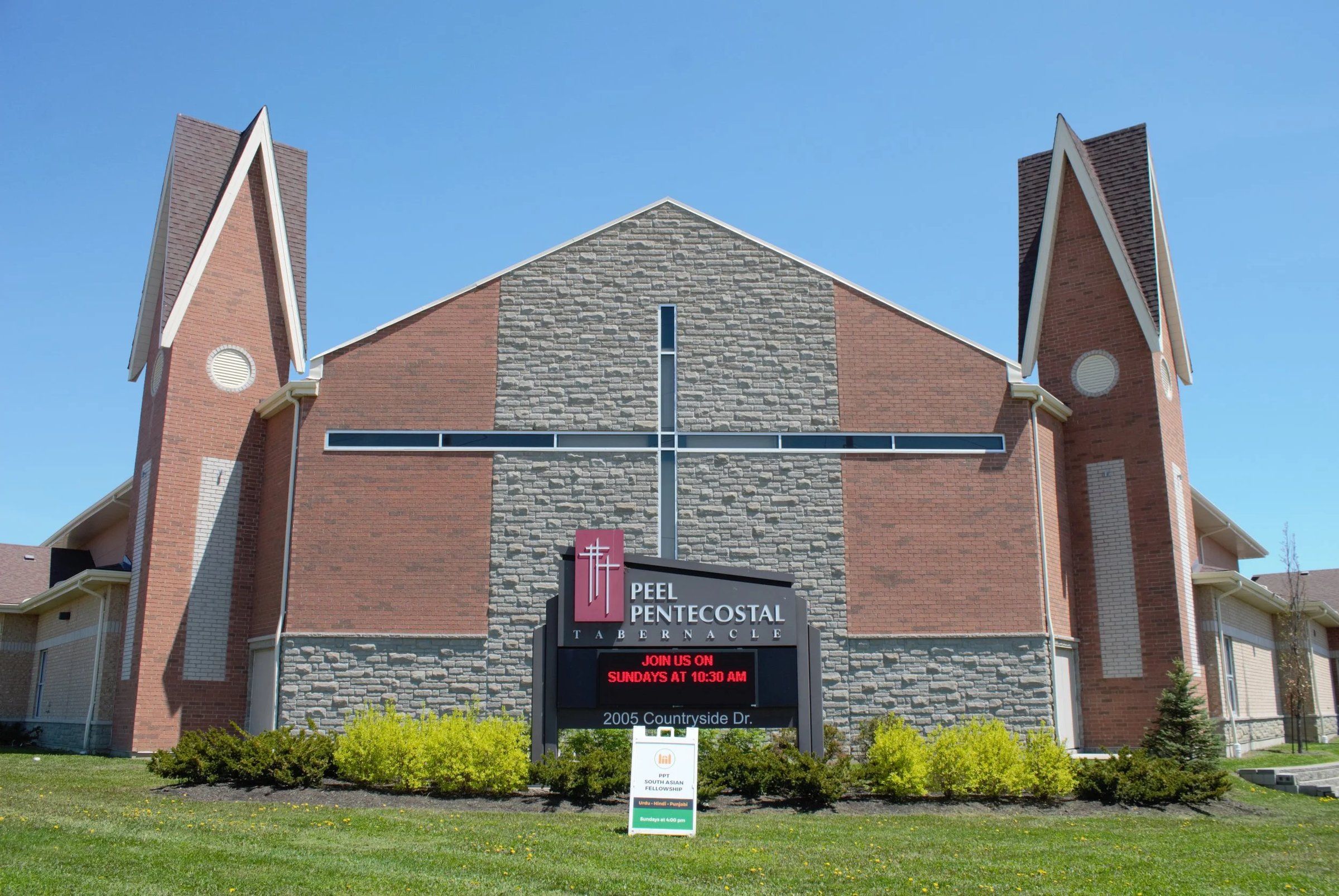 Peel Pentecostal Tabernacle, a brick and grey stone front with a tall cross set into it between two pointed towers, behind the church's sign