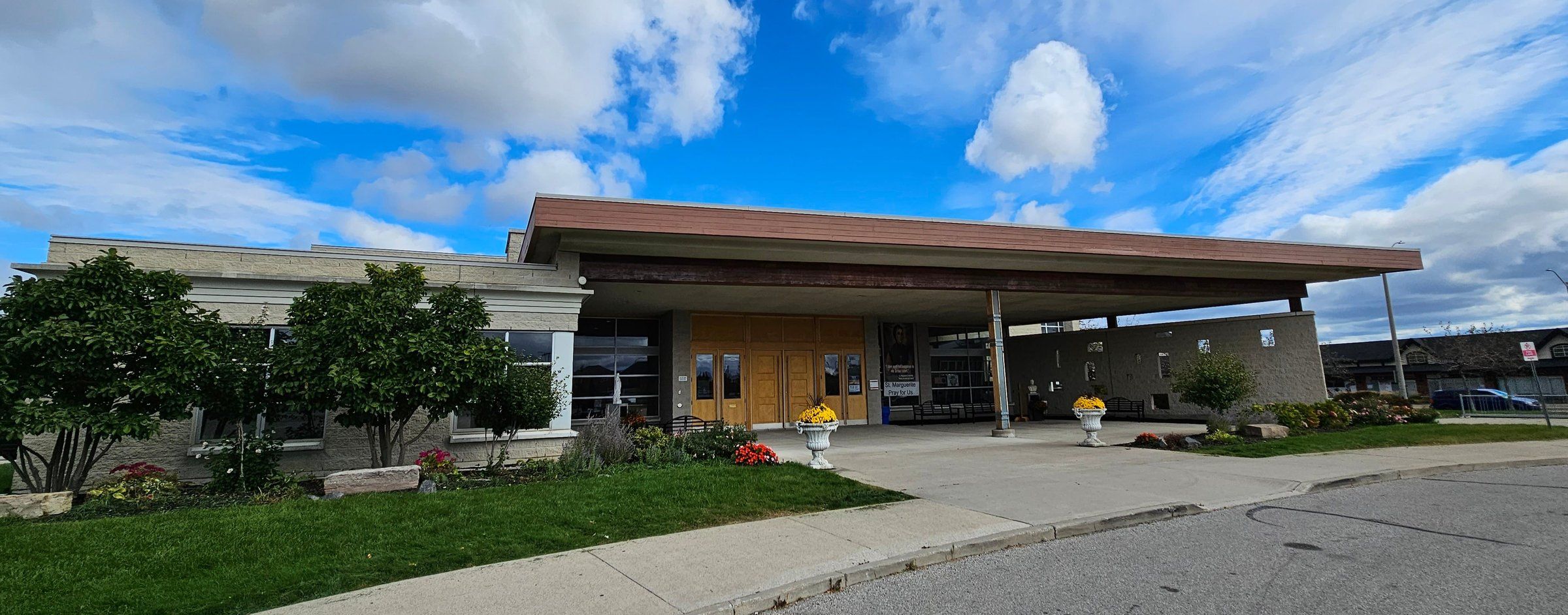 The entrance of St. Marguerite d'Youville Church under a long flat canopy, with trees and flower beds along the drive
